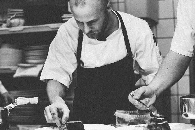 A black and white photograph shows two chefs in a professional kitchen setting, meticulously preparing food. The chef on the left, who is bald and has a short beard, is wearing a light-colored chef's jacket and a dark apron. He is focused on arranging small, dark-colored food items on a white plate with his hands. His expression is one of concentration. The chef on the right, partially visible in the frame, is holding a spoon in his right hand and appears to be adding sauce or liquid to another plate. He is also wearing a light-colored chef's jacket. In the background, out of focus, there are shelves with various kitchen items and what seems to be another person standing. The lighting in the kitchen is bright, highlighting the chefs and their work. The overall atmosphere suggests a busy and professional culinary environment.