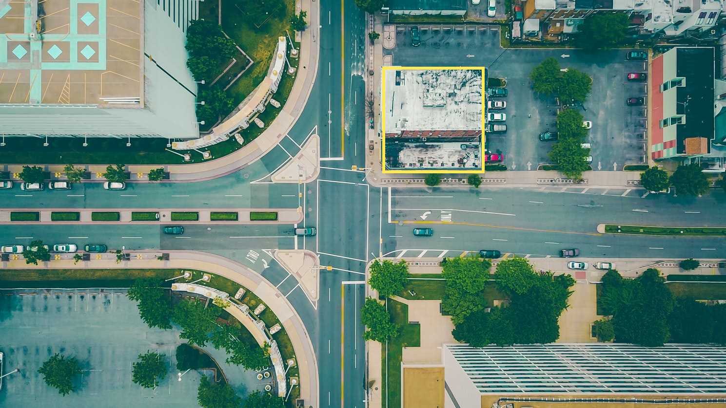 An aerial photograph taken directly above a four-way intersection in an urban environment. Two main roads intersect, each having multiple lanes for vehicular traffic. The road running horizontally across the image has a median strip with greenery. The road running vertically has a dedicated left-turn lane and a lane with a "ONLY" marking painted on the asphalt, indicating it's a one-way street for right turns only. Buildings of varying sizes and colors surround the intersection. On the top left, there's a large, light-colored building with geometric patterns on its roof. To the right of the vertical road, there's a long, narrow building with a white roof and several parked cars in front. Further to the right, a taller building with red and white vertical stripes is visible. Below the horizontal road, there are more buildings, including one with a light brown facade. Trees and patches of green vegetation are interspersed throughout the scene, adding natural elements to the urban landscape. The overall perspective is from a high vantage point, looking straight down at the intersection and its surroundings.
