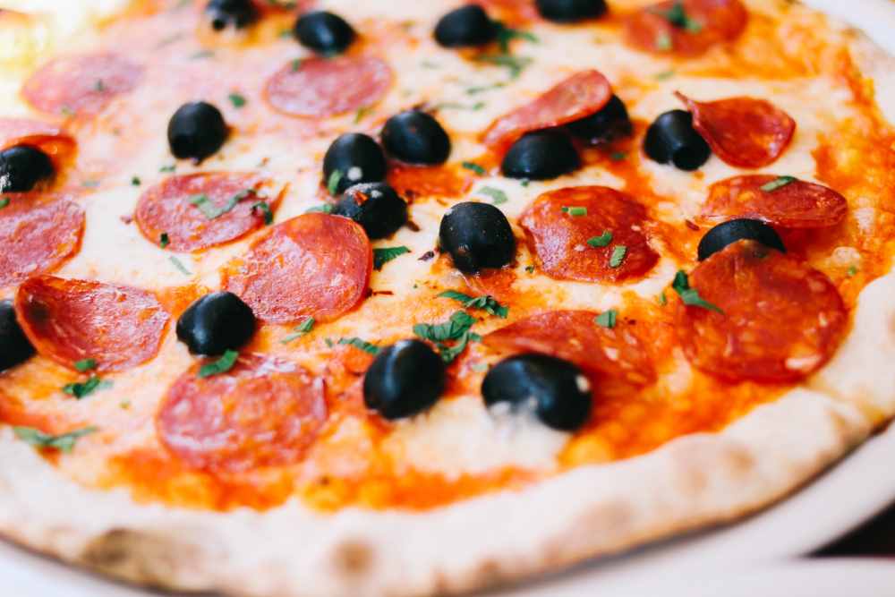 A close-up, high-angle shot showcases a delicious pizza with a golden-brown crust. The pizza is topped with melted white cheese, numerous round slices of bright red pepperoni, and several shiny black olives. The pepperoni slices are slightly curled at the edges, indicating they have been baked. Fresh green herbs, possibly oregano or parsley, are sprinkled sparsely across the pizza, adding a touch of color and visual texture. The crust is slightly raised and airy around the edges. The pizza appears to be freshly made and ready to be enjoyed. The lighting is bright and even, highlighting the vibrant colors and textures of the ingredients.
