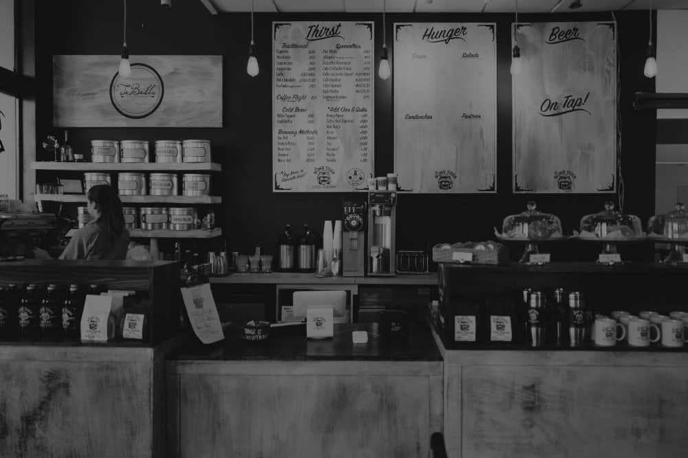 A black and white photograph captures the interior of a cozy restaurant or cafe. The scene is dimly lit by pendant lights hanging from the ceiling. On the back wall, three large menu boards are visible, listing options for "Thirst," "Hunger," and "Beer." Below these, a counter displays various items, including covered cakes or pastries under glass domes. To the left, a barista stands behind another counter, possibly preparing drinks. Shelves behind the barista are stocked with mugs and other supplies. In the foreground, a wooden counter extends across the frame, with paper menus or promotional materials placed on it. The overall ambiance is relaxed and inviting, suggesting a casual dining or social gathering spot. The black and white tones enhance the vintage or rustic feel of the establishment.